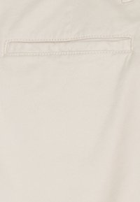 Tela de mezclilla beige que muestra un bolsillo trasero con una apertura oculta, con costuras tonales para un aspecto uniforme. La textura parece suave.