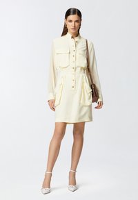 Abito camicia giallo a maniche corte con due tasche anteriori, chiusura a bottoni, vita cintrata e gonna plissettata. Abbinato a décolleté bianche con cinturino alla caviglia.
