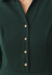 Vestido verde con frente abotonado, que presenta botones de madera y un cuello con solapa. La tela parece suave con una textura sutil.