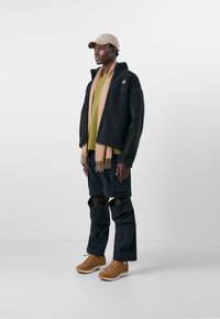 Schwarze Fleecejacke, grünes Hemd, beiger Schal, schwarze Hose mit Ausschnitten, tanfarbene Sneaker und beiger Hut. Einfaches Design und kontrastierende Farben.
