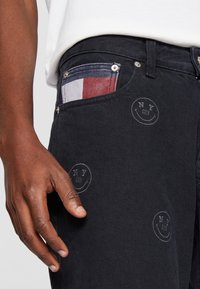 Dark denim jeans feature a patch pocket with a red, white, and blue checkered pattern, and embossed circular "NY" logos.