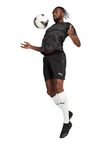 Schwarzes Kurzarmtrikot mit strukturiertem Muster, schwarze Shorts, schwarze Fußballschuhe und weiße Kniestrümpfe mit Logo, Spieler jongliert mit einem Fußball.