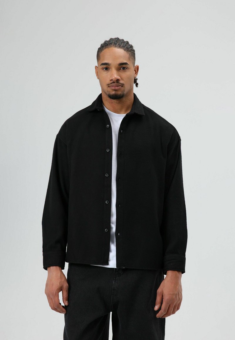 Zwarte overhemd met knopen en een kraag, gemaakt van een gestructureerde stof, met lange mouwen en een rechte snit. Gedragen over een wit T-shirt.