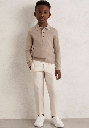 Polo shirt beige à manches longues avec trois boutons, associé à un pantalon blanc ajusté et des baskets blanches. Texture douce et design ajusté.
