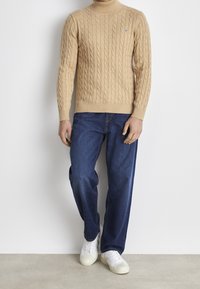 Helles kabelgestricktes Rollkragensweatshirt, blaue Jeans und weiße Sneakers. Das Sweatshirt hat gerippte Bündchen und einen Saum sowie ein kleines Logo auf der Brust.