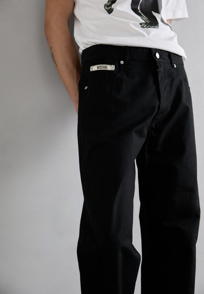 Svarta denimjeans med avslappnad passform, synlig sömnad och metalldetaljer. Märket på midjebandet säger "MOSCHINO."