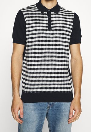 Homme portant un polo noir à manches courtes avec un panneau avant à carreaux noir et blanc, associé à un jean en denim bleu clair.