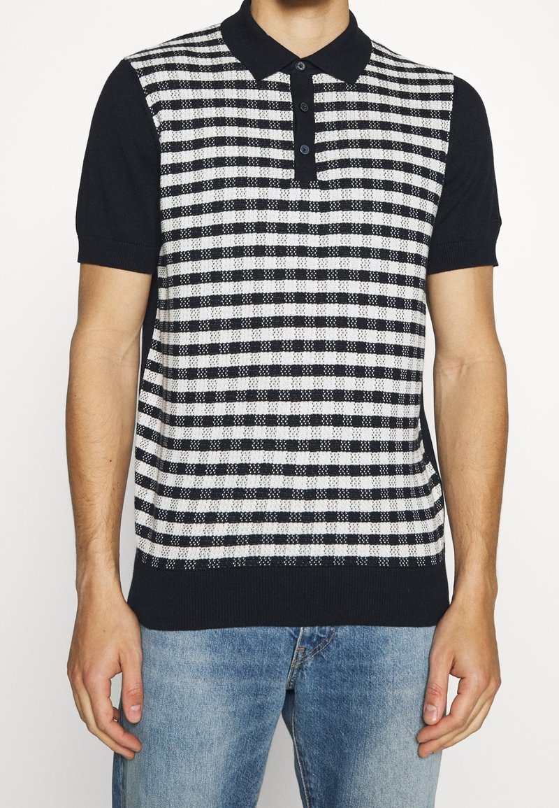 Homme portant un polo noir à manches courtes avec un panneau avant à carreaux noir et blanc, associé à un jean en denim bleu clair.