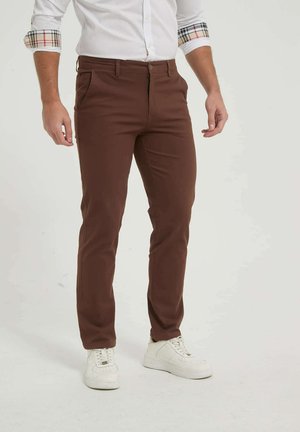 Homme portant un pantalon marron ajusté, des baskets blanches et une chemise blanche aux manches retroussées laissant apparaître des revers à carreaux, debout sur un fond blanc.