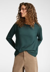 Suéter de manga larga verde oscuro con un corte relajado, cuello redondo y textura suave. Combinado con pantalones beige claros. Diseño simple y versátil.