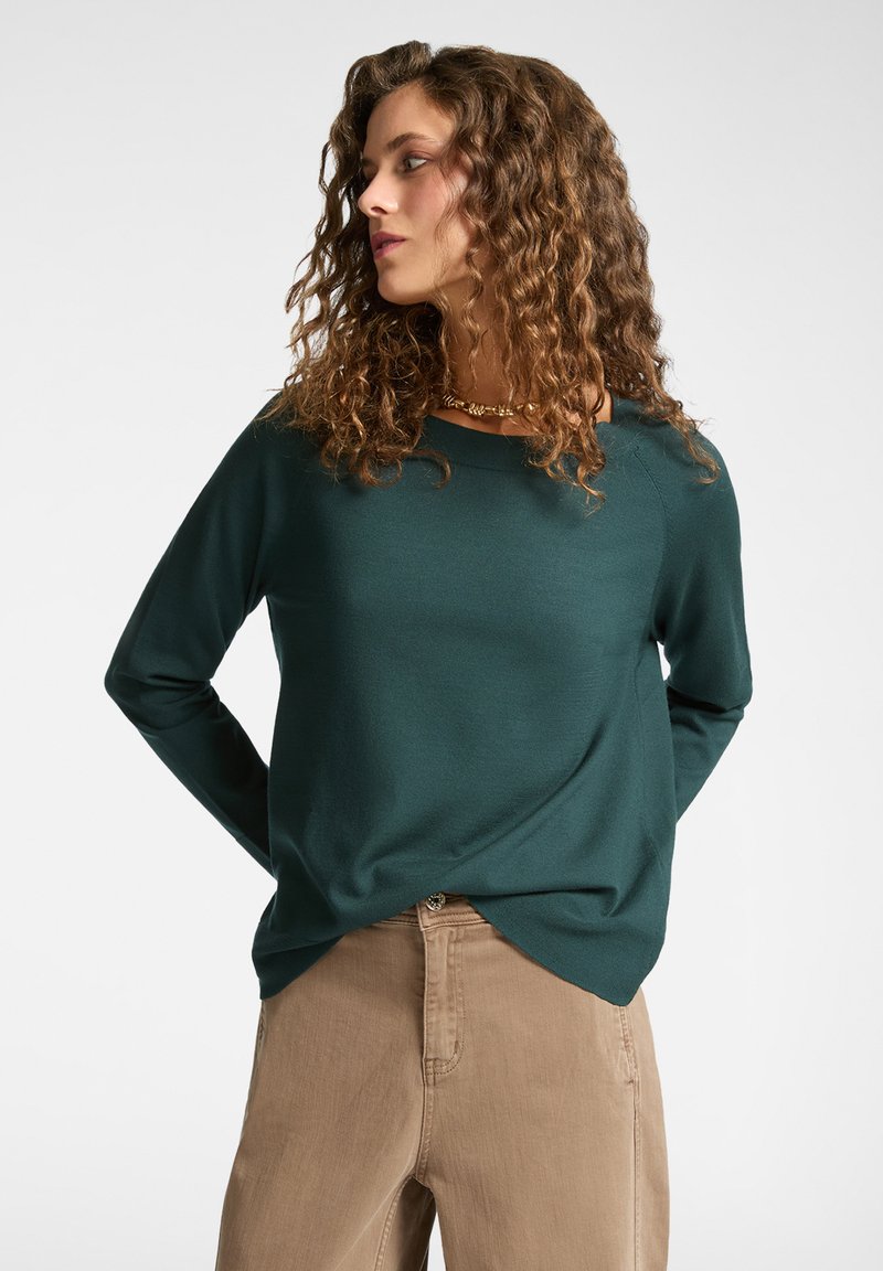 Suéter de manga larga verde oscuro con un corte relajado, cuello redondo y textura suave. Combinado con pantalones beige claros. Diseño simple y versátil.