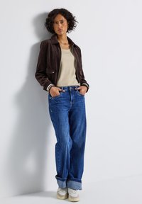 Brązowa zamszowa bomberka z białymi paskami na mankietach, beżowy top, niebieskie szerokie dżinsy i białe sneakersy z jasnymi akcentami.