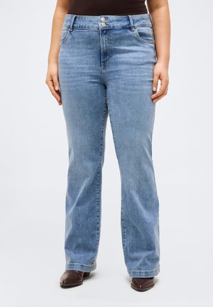 Femme portant un jean bleu clair taille haute avec fermeture à double bouton, poches avant et chaussures marron à bouts pointus, debout les mains détendues.