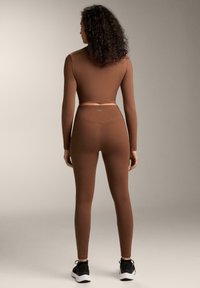 Brązowa bluzka z długim rękawem i dopasowane legginsy z gładkiego materiału; posiadają bezszwową konstrukcję i elastyczną talię. Połączone z czarnymi trampkami.