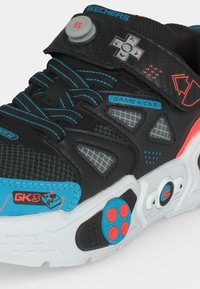 Μαύρο και μπλε sneaker Skechers με σχέδια κουμπιών χειριστηρίου παιχνιδιών και ετικέτα "Game Kicks" στο πλάι, με μπλε και κόκκινες λεπτομέρειες.