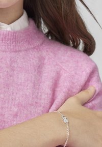 Maglione rosa con scollatura rotonda abbinato a un braccialetto in argento con un piccolo charm a forma di foglia su un polso di carnagione chiara.
