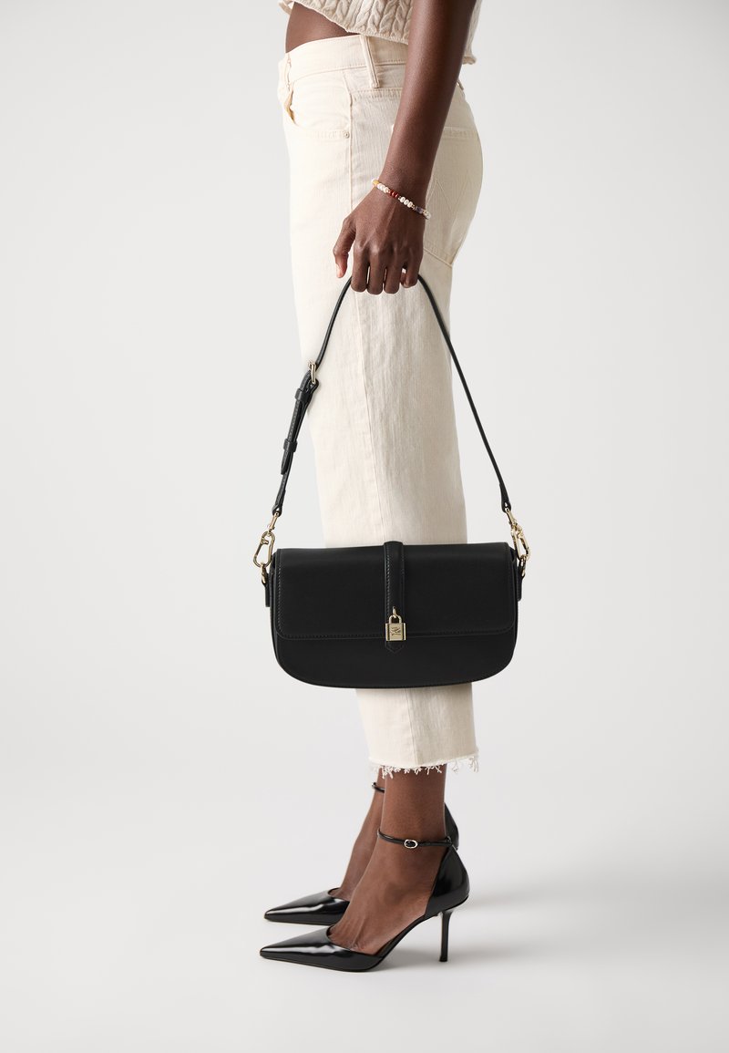 Black leather handbag with a top flap, gold hardware, side strap, and structured design. Worn by a person in light-colored pants and heels.