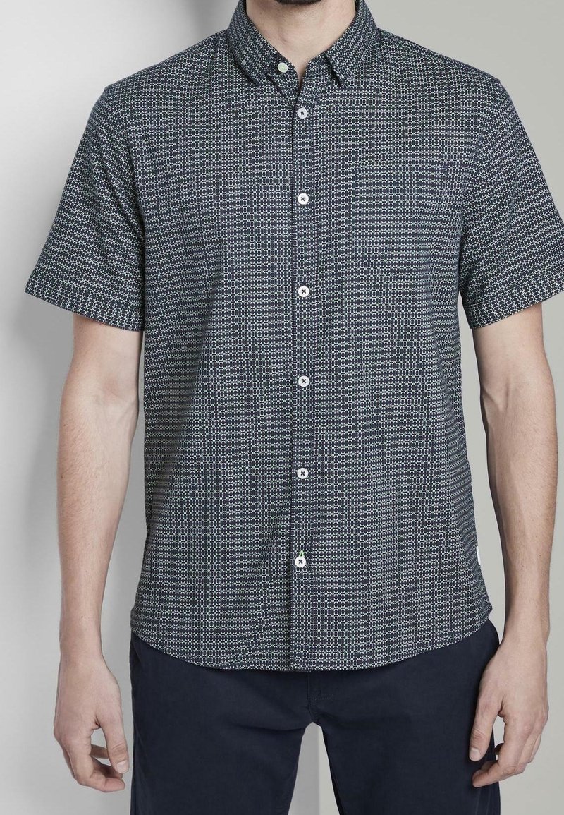 Homme portant une chemise bleu marine à motifs à manches courtes avec col et pantalon foncé, debout devant un fond uni.