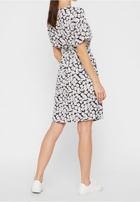 Woman wearing a knee-length navy dress with white floral print and white sneakers, standing with back facing the camera.