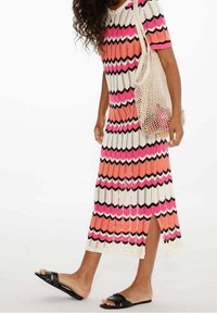 Personne portant une robe colorée à motif zigzag avec une fente sur le côté, tenant un sac en filet avec des fruits, et portant des sandales noires sans lacets.