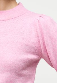 Κοντινό πλάνο ενός ατόμου που φοράει ένα απαλό ροζ πλεκτό πουλόβερ με ψηλό ριμπ πλεκτό γιακά και φουσκωτές λεπτομέρειες στους ώμους.