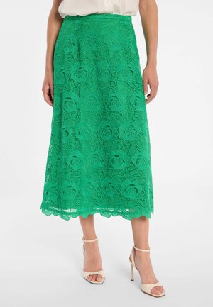 Femme portant une jupe midi en dentelle verte à motifs floraux et des sandales à talons hauts beiges, debout devant un fond blanc.