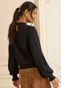 Fekete, puffos ujjú felső hátul kulcstartó kivágással, bordázott mandzsettákkal és sima textúrával, barna velúr szoknyával párosítva.