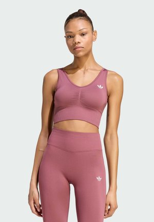 Jonge vrouw die een bijpassende mauvekleurige sportieve crop top en high-waisted leggings draagt, staand tegen een eenvoudige lichte achtergrond.