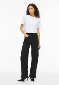 Balti trumpomis rankovėmis marškinėliai, derinami su juodomis kelnėmis platėjančiomis nuo juosmens ir juodais smailiais aukštakulniais, pabrėžiantys švarią, modernią siluetą.