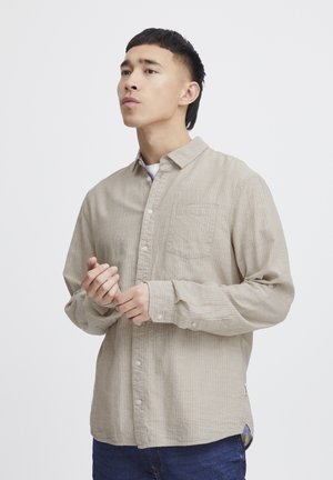 Beige long-sleeve button-up shirt with a subtle striped pattern, single chest pocket, and a curved hem. Worn over a white shirt.
