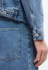 Κοντινό πλάνο ενός ατόμου που φοράει μπλε τζιν μπουφάν και τζιν παντελόνι με μεταλλικά κουμπιά, εστιάζοντας στην πίσω τσέπη και τις λεπτομέρειες στις μανσέτες.