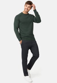 Un pull en maille vert foncé à col rond avec un ourlet côtelé, associé à un pantalon sombre ajusté et des baskets blanches, présentant une texture lisse.