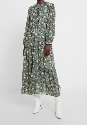 Kobieta ubrana w długą sukienkę z długim rękawem i sięgającą kostek, z zielonym wzorem liści i białych kwiatów, w białych botkach na obcasie, stojąca na białej podłodze.