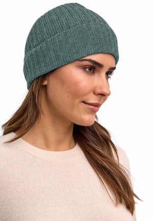 Vrouw met lang bruin haar, draagt een groen geribbelde gebreide muts en een lichtbeige trui, kijkt naar rechts tegen een witte achtergrond.