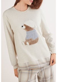 Sudadera beige claro con diseño de oso con un jersey azul estampado, mangas largas y cuello redondeado. Pantalones de cuadros abajo.