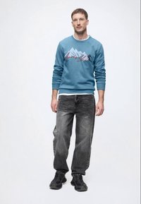 Hanorac albastru cu grafică a unui munte și text, asociat cu blugi gri închis și adidași negri. Siluetă simplă, relaxată și stil casual.