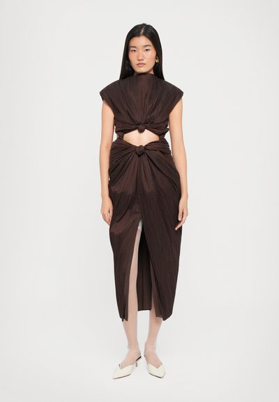 Robe plissée marron avec un col montant, des détails noués à la taille et aux épaules, et une fente jusqu'à la cuisse. Complétée par des chaussures de couleur claire.