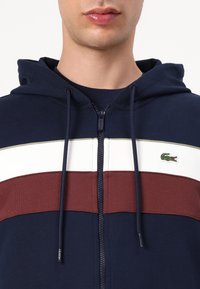 Sweatshirt navy com fecho, com riscas horizontais brancas e bordô, apresentando um pequeno logótipo de crocodilo verde e cordões na capuz.