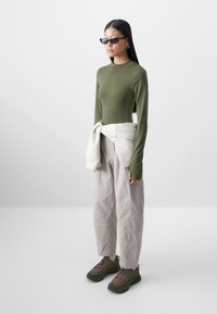 Haut à manches longues vert olive, pantalon taille haute gris clair et chaussures chunky brun-vert ; lunettes de soleil noires et sweat-shirt blanc noué autour de la taille.