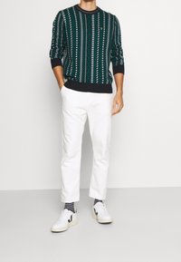 Suéter verde de punto con patrones geométricos en blanco y negro, combinado con pantalones blancos y zapatillas blancas con acentos negros.
