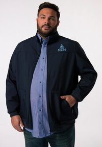 Giubbotto softshell blu navy con colletto alto e zip frontale, dotato di due tasche e un logo blu sul petto. Indossato sopra una camicia azzurra chiara.