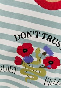 Pruhovaná světle modrá a bílá košile s krátkým rukávem, červeným výstřihem, na kterém je výšivka s textem, květinami a motivem hada.