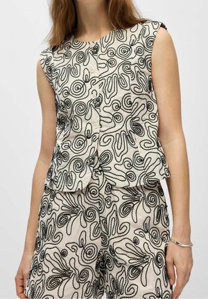 Femme portant un haut sans manches boutonné et un short assorti avec des motifs tourbillonnants abstraits noirs sur un tissu clair, debout les mains détendues.