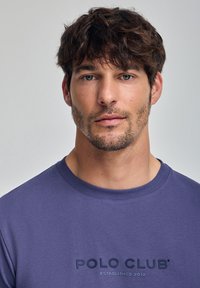 Lila crew-neck t-shirt med broderad text "POLO CLUB" och etableringsdatum; bomullsmaterial med en mjuk textur.