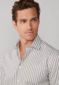 Biała i jasno szara koszula na guziki w paski z kołnierzykiem, o delikatnym połysku i teksturowanej tkaninie. Widoczne są gładkie guziki.