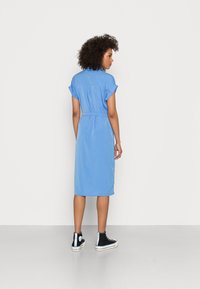 Robe bleue jusqu'aux genoux avec des manches courtes et retroussées, taille ceinturée et texture lisse. Assortie à des baskets montantes noires avec des accents à motifs.
