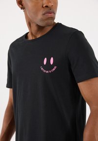 Svart bomullst-skjorte med et frontmotiv i rosa med et smilende ansikt og teksten "EN BEDRE MEG KOMMER." Korte ermer, rund hals.