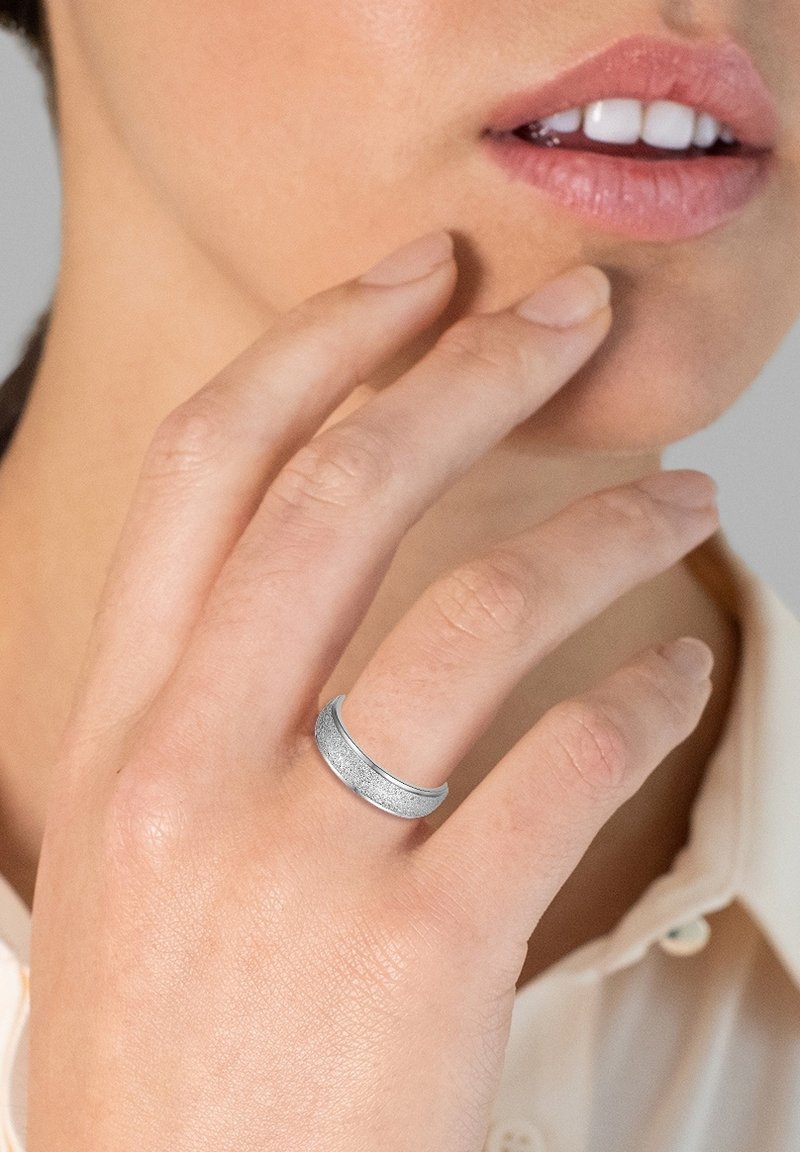 Anillo de plata con un acabado texturizado, que presenta un borde superior curvado y suave. Lleva puesto en la yema de un dedo, sobre un fondo de tono de piel.
