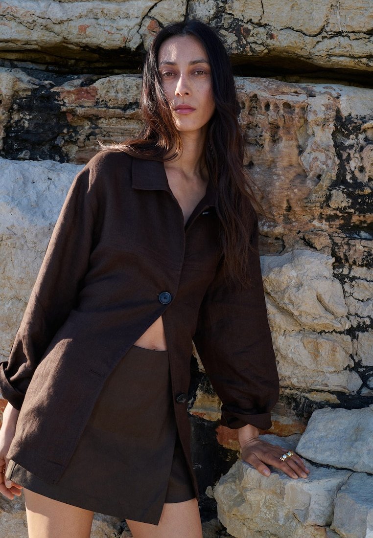 Femme aux longs cheveux foncés portant une veste et une jupe marron foncé, appuyée contre des rochers texturés à la lumière naturelle.