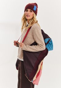 Vícebarevná péřová bunda v barvách mahagonové, červené a modré. Naražená přes béžový svetr se vzorem copánku, s červeným popruhem a modrými akcenty.